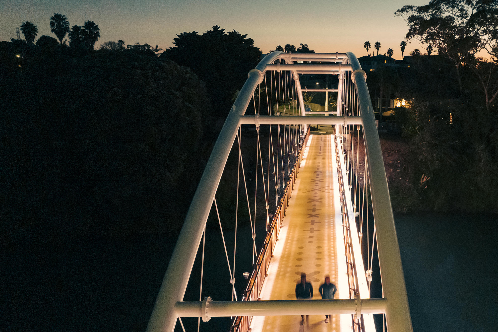 Illuminated white arch bridge at dusk. Its pathway glows with warm light as two figures walk in the distance. Palm trees and hills are silhouetted against the twilight sky