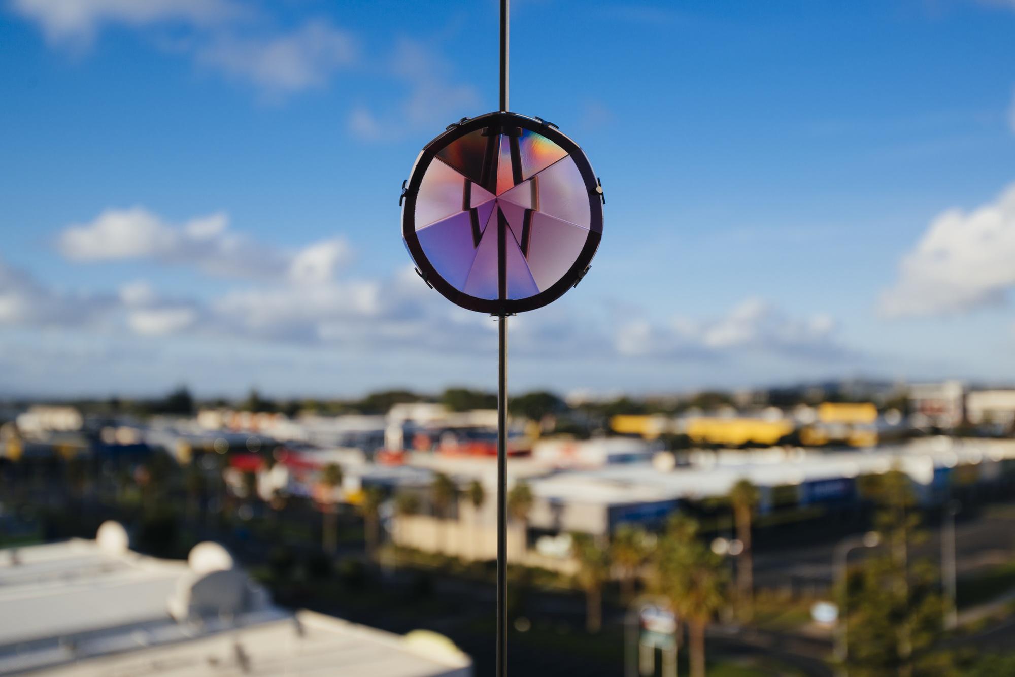 A faceted prism reflecting a colourful spectrum, suspended against a bright blue sky with scattered clouds, overlooking a blurred urban landscape