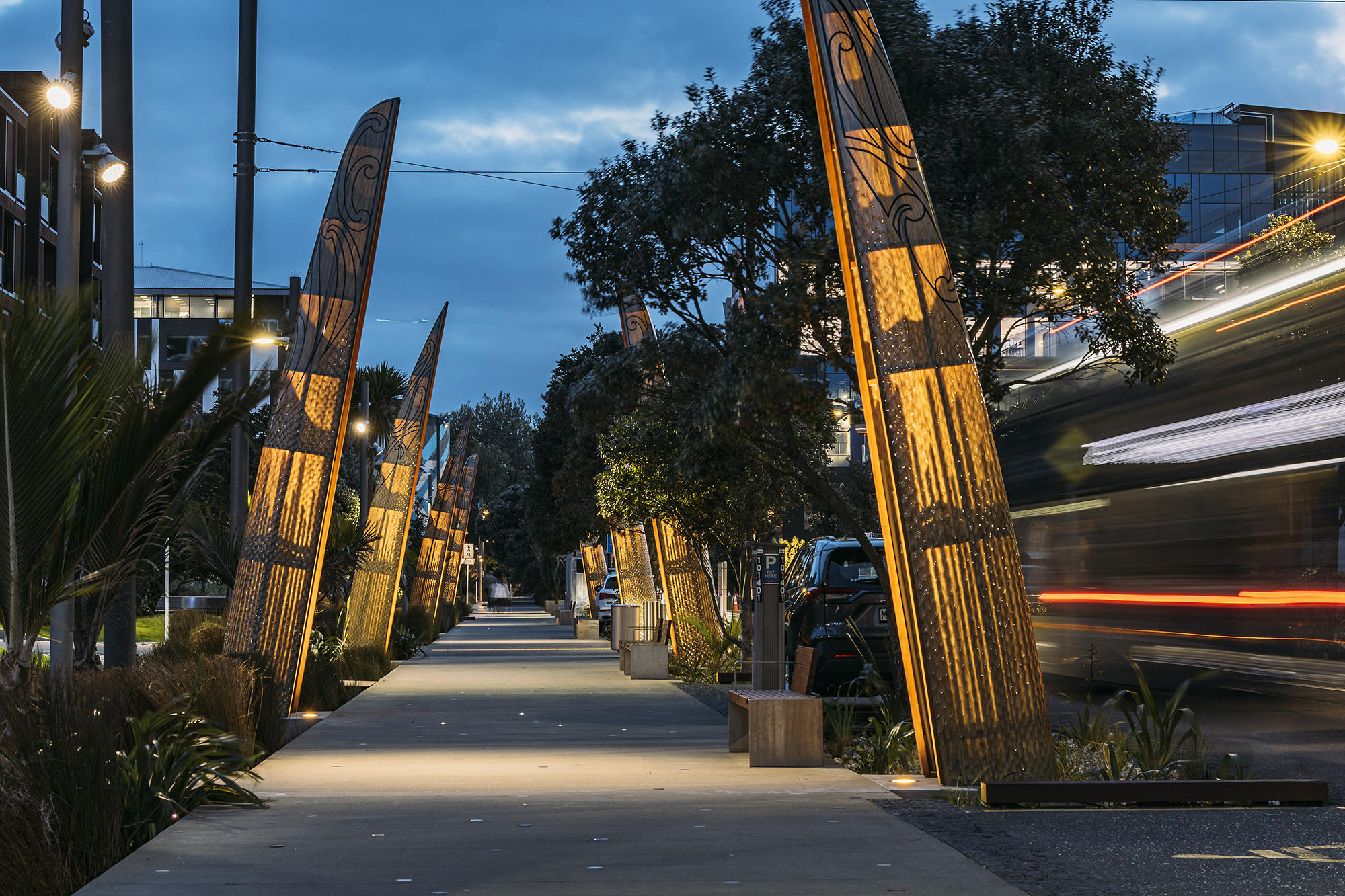 Pedestrian pathway at dusk with tall carved wooden sculptures lit from below, trees and plants on both sides, blurred vehicle with light trails on the right, and buildings in the background under a cloudy sky.