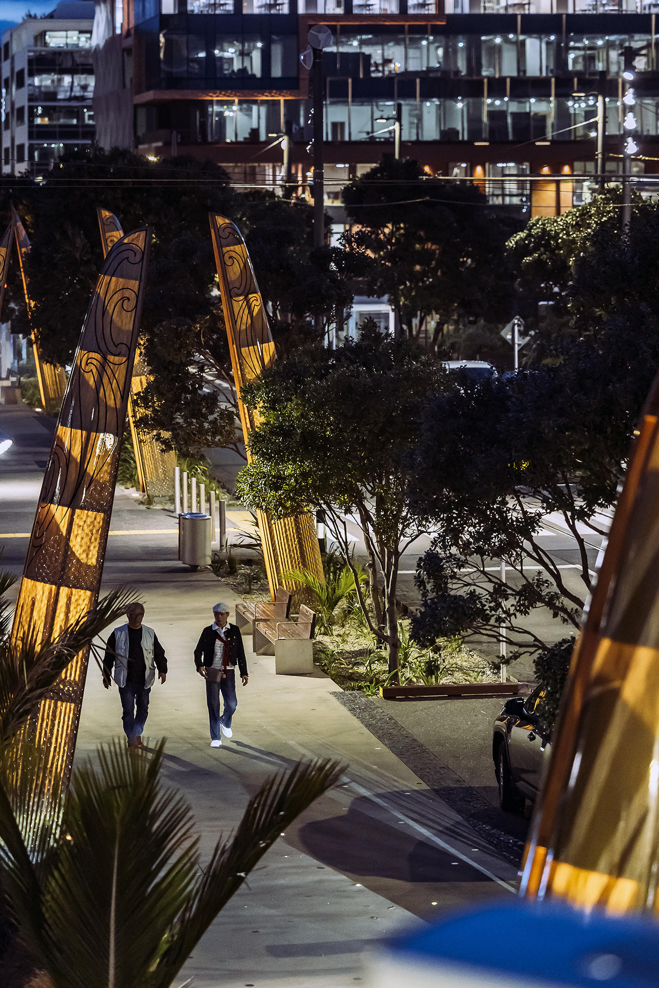 Nighttime urban scene with two people walking along a well-lit pathway lined with tall carved wooden structures. Modern multi-storey buildings with glass windows are visible in the background, and trees and greenery frame the area.