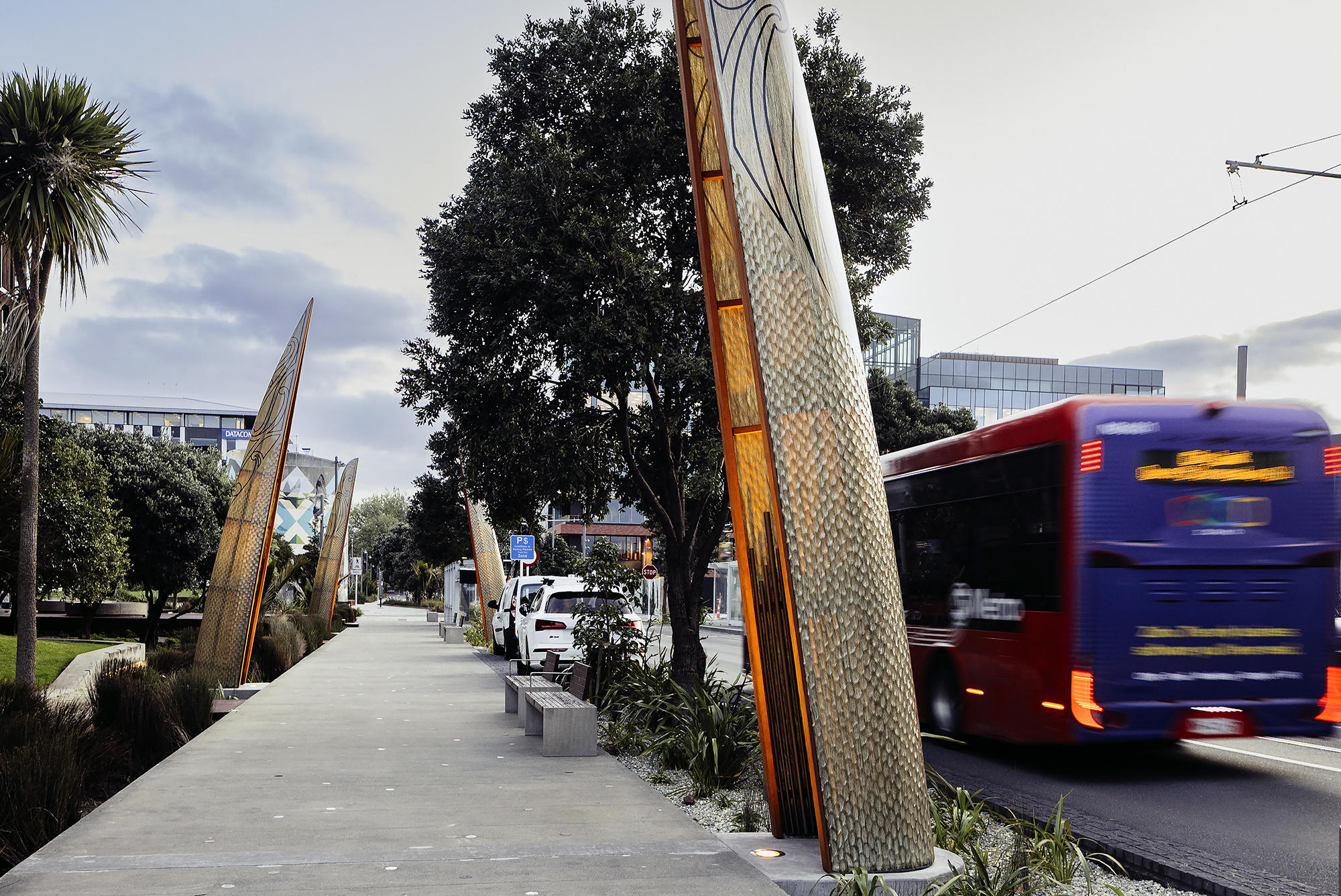 Pathway lined with tall, patterned metal structures resembling waka hulls, bordered by trees and plants, with a red bus driving on the adjacent road under a partly cloudy sky.