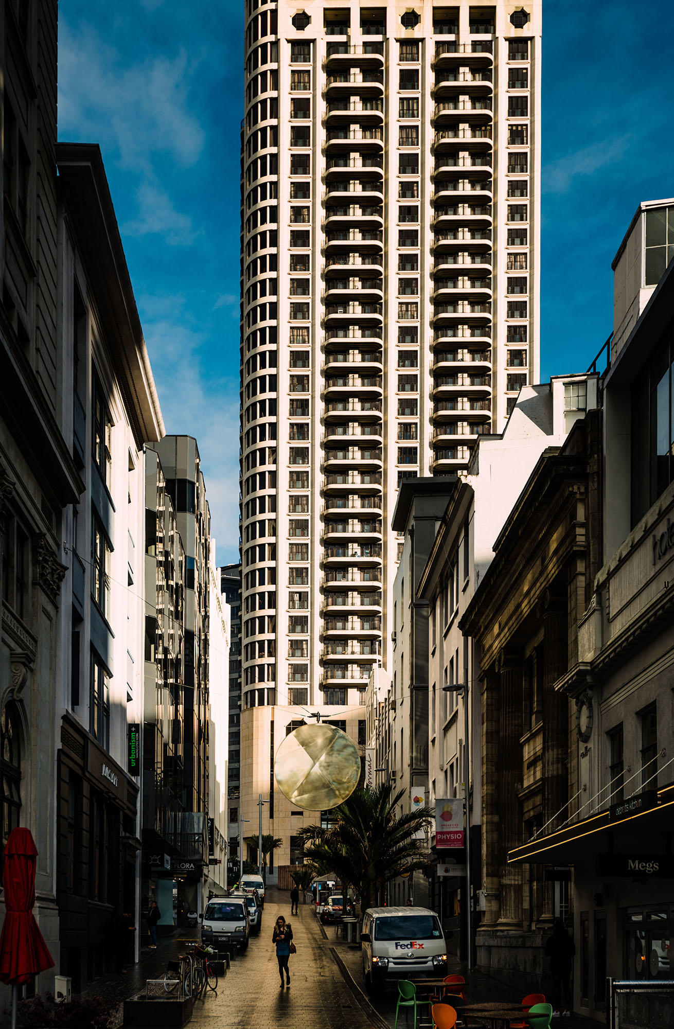 A narrow lane with a large round mirror suspended above, a tall skyscraper behind