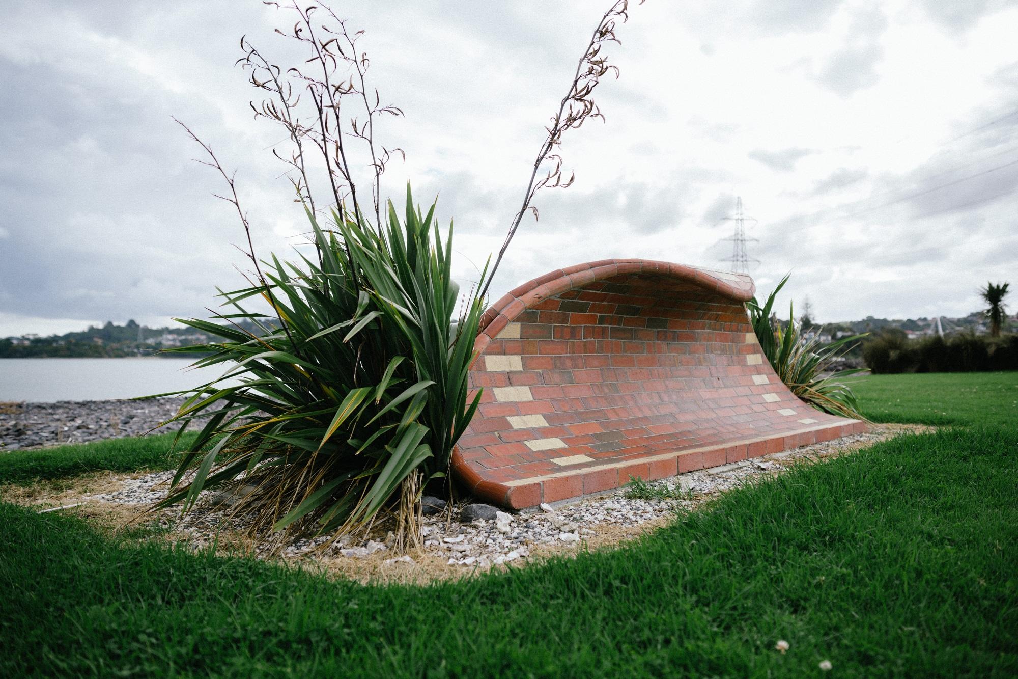 Side view of a brick sculpture in the shape of a wave with a flax bush in the foreground