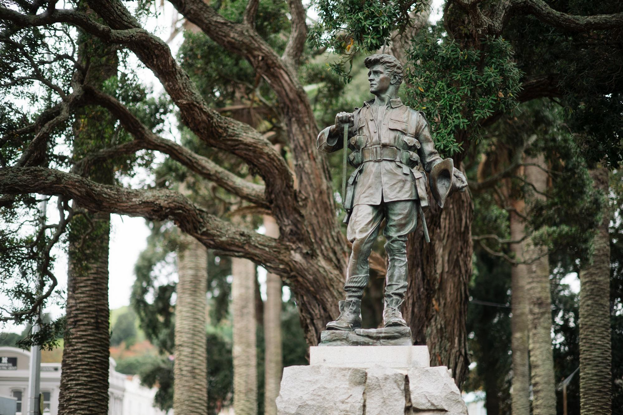 A bronze statue of a soldier looking left stands on a plinth with tree branches behind